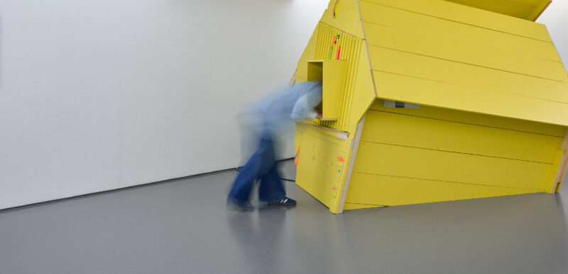 Hosting Infrastructure is an installation by Fergus Tibbs. The image shows an installation view with a blurred human figure leaning into the window of a large yellow house-shaped structure.